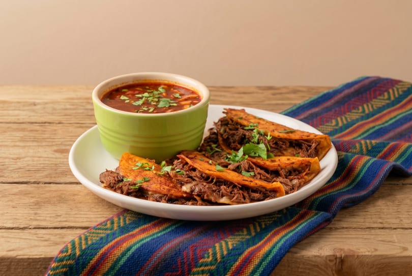 Three birria tacos with cilantro on a white plate, next to a green bowl of consommé, on a wooden table.