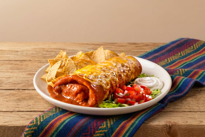 A cheesy, saucy burrito with tortilla chips, pico de gallo, and sour cream on a wooden table.